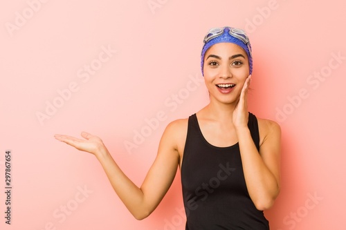 Young arab swimmer woman isolated holds copy space on a palm, keep hand over cheek. Amazed and delighted.