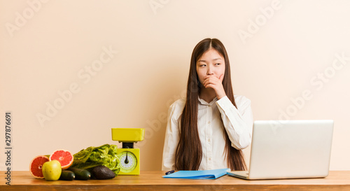 Young nutritionist chinese woman working with her laptop thoughtful looking to a copy space covering mouth with hand.