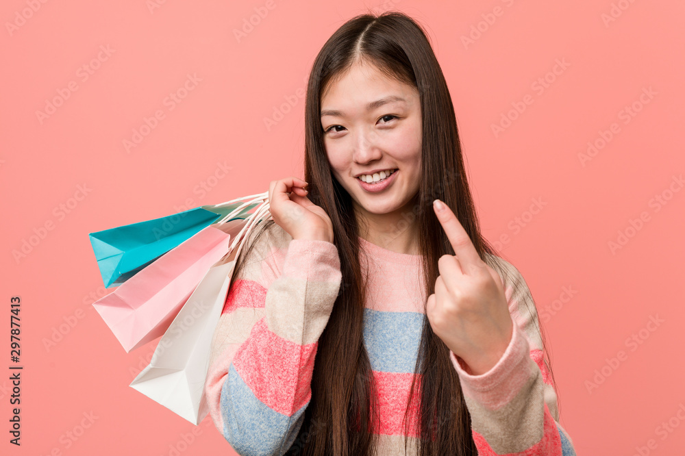 Young asian woman holding a shopping bag pointing with finger at you as if inviting come closer. Young asian woman holding a shopping bag pointing with finger at you as if inviting come closer.
