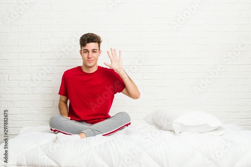 Young teenager student man on the bed smiling cheerful showing number five with fingers.