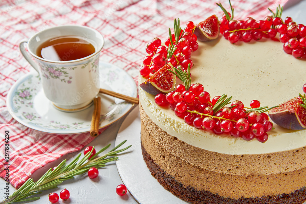 Round homemade cake decorated with red currants, figs, cinnamon and rosemary with tablecloth, Cup and saucer on a light background concrete table.