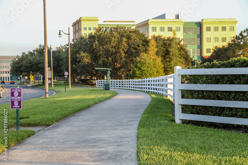 Wide white, curved fence, beside a sidewalk with grass, in the background the beautiful landscape of plants and trees, in Celebration Street, Orlando, Florida, USA.