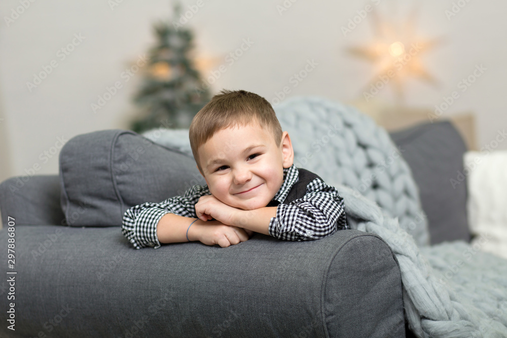 A little boy lies on a sofa covered with a gray knitted blanket and smiles, against the background of Christmas decor and a Christmas tree A little boy lies on a sofa covered with a gray knitted blanket and smiles, against the background of Christmas decor and a Christmas tree