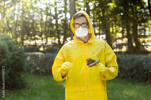 Man yellow protective suit, glasses and respirator holds dry leaves in one hand. Portrait of man on the background of blurred trees