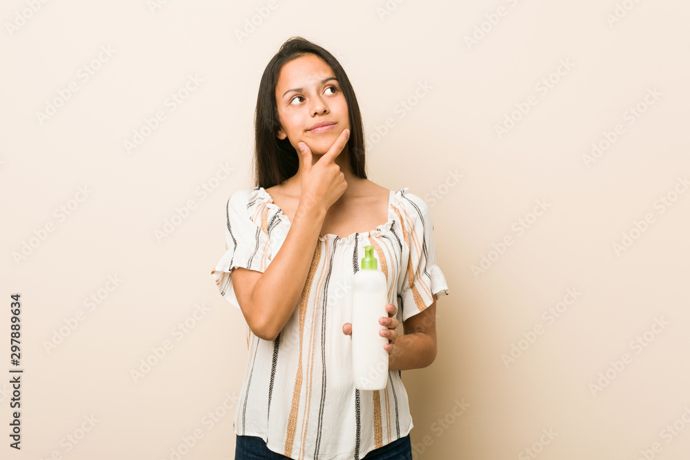 Young hispanic woman holding a cream bottle looking sideways with doubtful and skeptical expression. Young hispanic woman holding a cream bottle looking sideways with doubtful and skeptical expression.