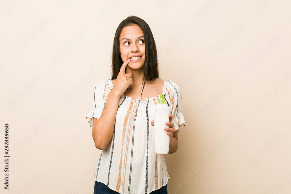 Young hispanic woman holding a cream bottle relaxed thinking about something looking at a copy space. Young hispanic woman holding a cream bottle relaxed thinking about something looking at a copy space.