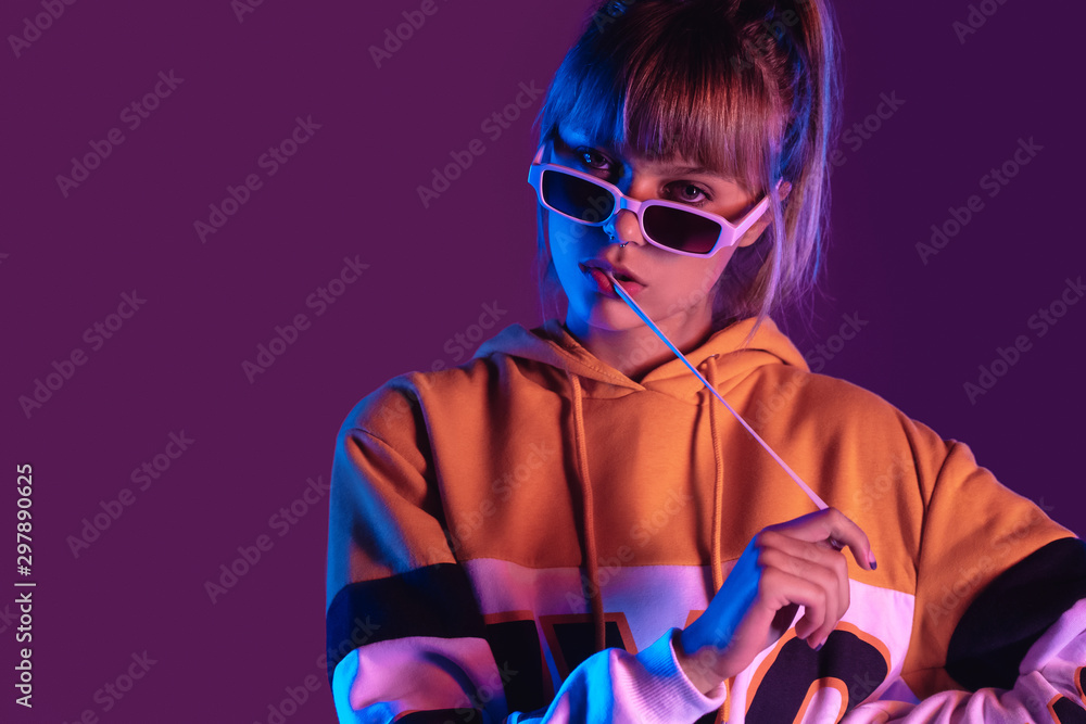 Stylish pretty young 20s fashion teen girl model wear glasses chew bubble gum look at camera stand at purple studio background, igen teenager with bubblegum in trendy night 80s party light portrait - obrazy, fototapety, plakaty 