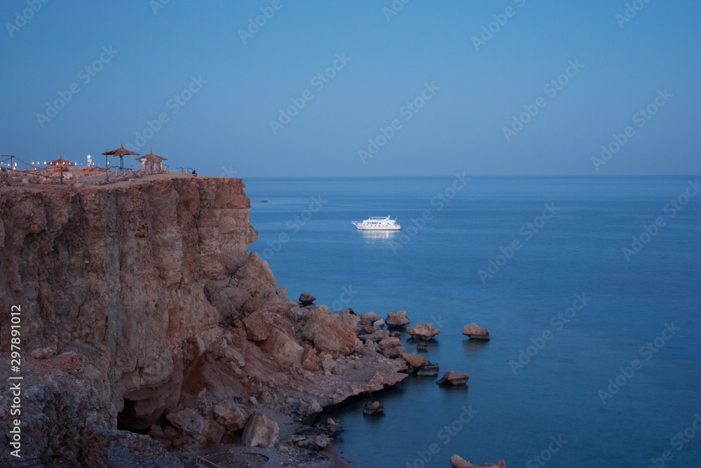 evening sea view with cliff cape in pink light, white boat in the sea and blue horizon