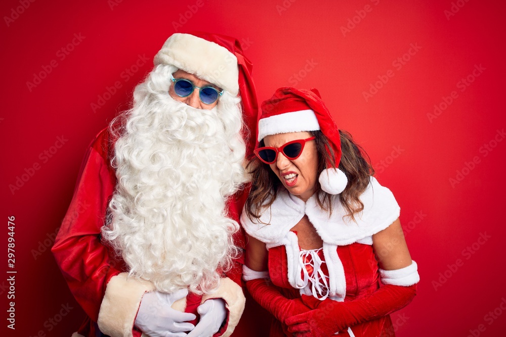 Middle age couple wearing Santa costume and sunglasses over isolated red background with hand on stomach because indigestion, painful illness feeling unwell. Ache concept. Middle age couple wearing Santa costume and sunglasses over isolated red background with hand on stomach because indigestion, painful illness feeling unwell. Ache concept.