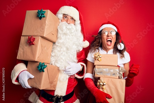 Middle age couple wearing Santa costume holding tower of gifts over isolated red background angry and mad screaming frustrated and furious, shouting with anger. Rage and aggressive concept.