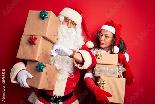Middle age couple wearing Santa costume holding tower of gifts over isolated red background Checking the time on wrist watch, relaxed and confident