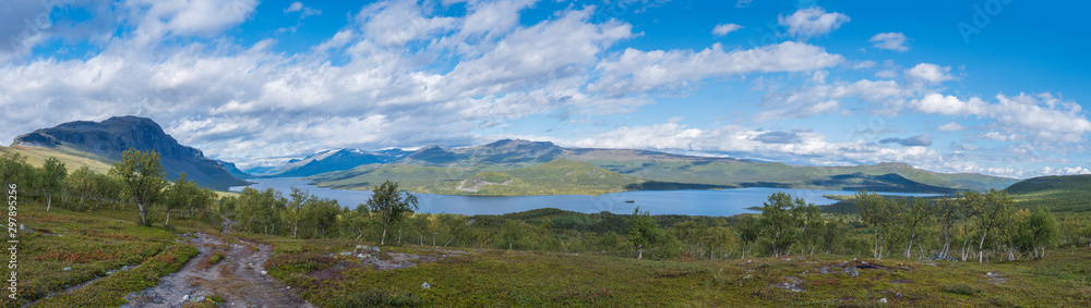 Fototapeta premium Panoramic landscape with beautiful river Lulealven, snow capped mountain, birch tree and footpath of Kungsleden hiking trail near Saltoluokta, north of Sweden, Lapland wild nature. Summer blue sky