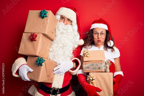 Middle age couple wearing Santa costume holding tower of gifts over isolated red background puffing cheeks with funny face. Mouth inflated with air, crazy expression.