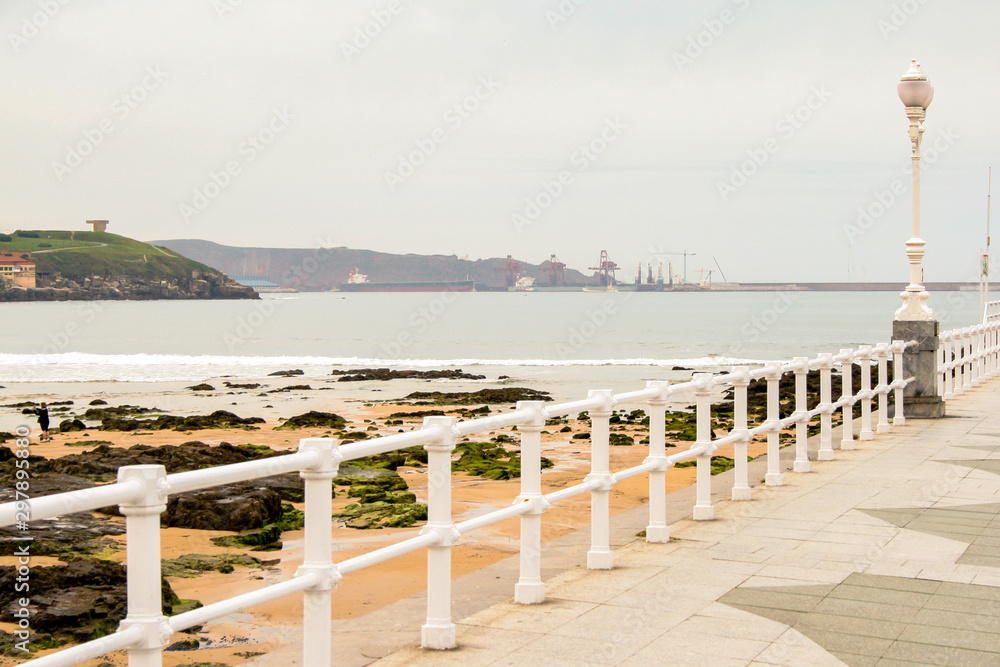 Promenade with white fences near a beach and rocks. Shipyard and oil tankers in the background on a cloudy day