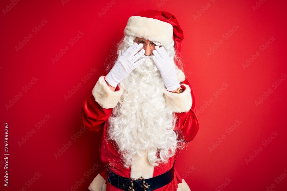 Middle age handsome man wearing Santa costume standing over isolated red background rubbing eyes for fatigue and headache, sleepy and tired expression. Vision problem