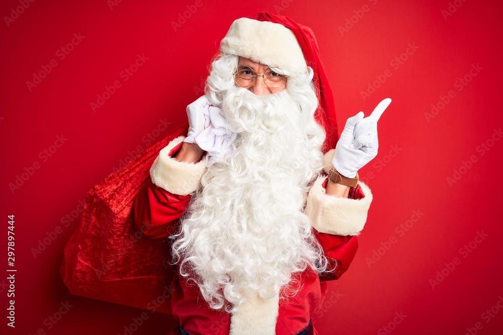 Middle age man wearing Santa costume holding sack with gifts over isolated red background very happy pointing with hand and finger to the side
