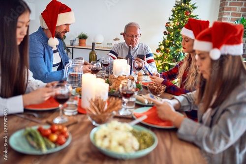 Beautiful family wearing santa claus hat meeting smiling happy and confident. Eating roasted turkey using smartphone celebrating Christmas at home