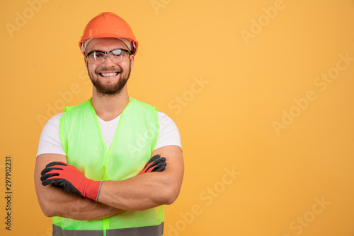 Positive happy foreman in workwear, keeps arms folded, wears orange helmet, receives new task from boss, glad to be promoted, stands against yellow background, blank free space. Repairman indoor