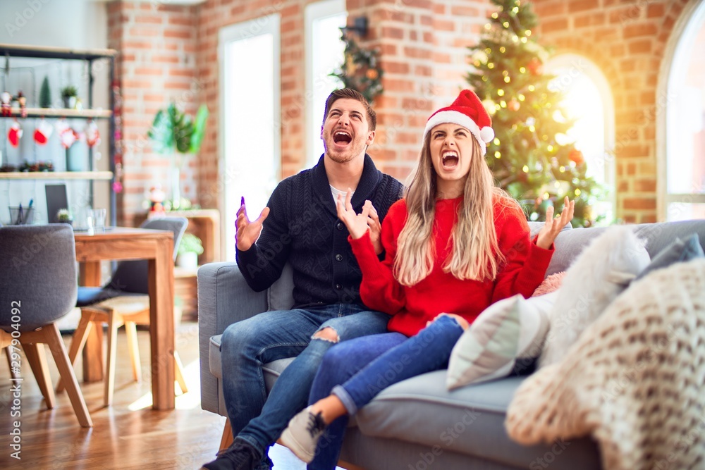 © Krakenimages.com - Young couple wearing santa claus hat sitting on the sofa around christmas tree at home crazy and mad shouting and yelling with aggressive expression and arms raised. Frustration concept.