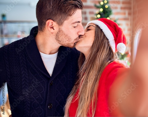 Young beautiful couple smiling happy and confident. Standing and kissing make selfie by camera around christmas tree at home