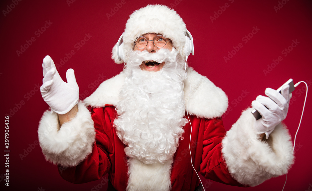 Fototapeta premium Carol of the bells. Authentic Santa Claus is posing with a joyful face expression, holding a smartphone in his left hand and listening to music on the headphones.