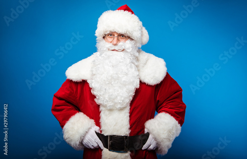Ho-ho-ho! Real Santa is posing on a blue background with both of his hands on a belt, smiling and looking at the camera.