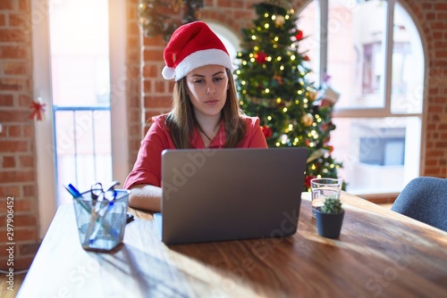 Beautiful woman sitting at the table working with laptop wearing santa claus hat at christmas with serious expression on face. Simple and natural looking at the camera.