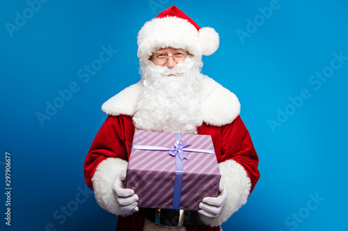 Mystery in a box. Close up photo of authentic Santa Claus, who is holding a violet striped present box with a purple ribbon right in front of him, smiling and looking gently at the camera.