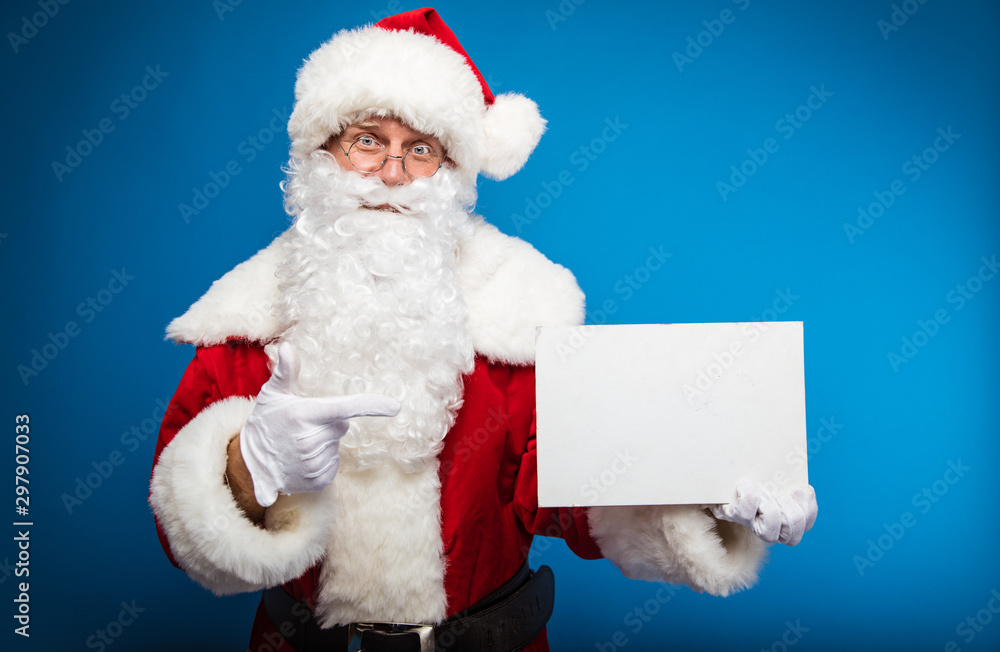 Naklejka premium My wishes to you. Santa Claus pointing with his right index finger at the blank postcard with copyspace on it, which he holds in his left hand, while looking at the camera.