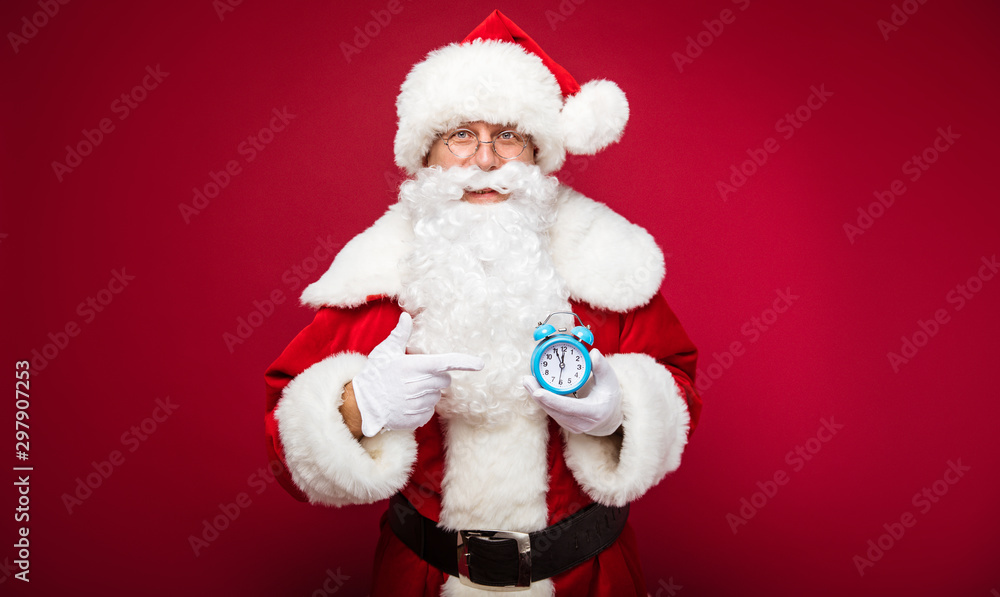 Look! Happy Santa pointing with his right index finger at the alarm clock, which shows five minutes to twelve, in his left hand and looking at the camera. Look! Happy Santa pointing with his right index finger at the alarm clock, which shows five minutes to twelve, in his left hand and looking at the camera.