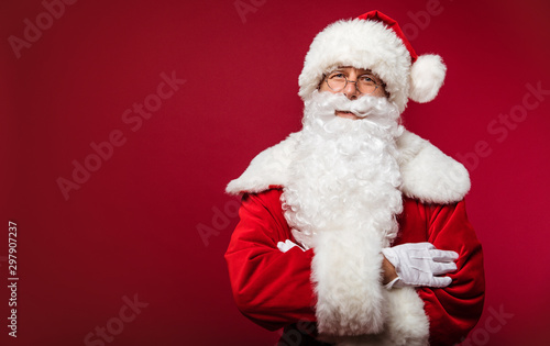Merry Christmas. Authentic Santa Claus with a long white beard and round-shaped glasses is standing with folded arms on a red background.