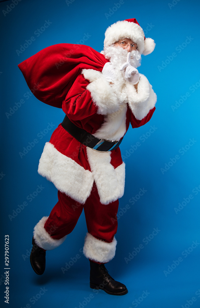 Don’t give me out! Full-length photo of Santa Claus, running to the right with a sack of presents on his right shoulder, showing a shush gesture with his left hand, and looking to the camera. Don’t give me out! Full-length photo of Santa Claus, running to the right with a sack of presents on his right shoulder, showing a shush gesture with his left hand, and looking to the camera.
