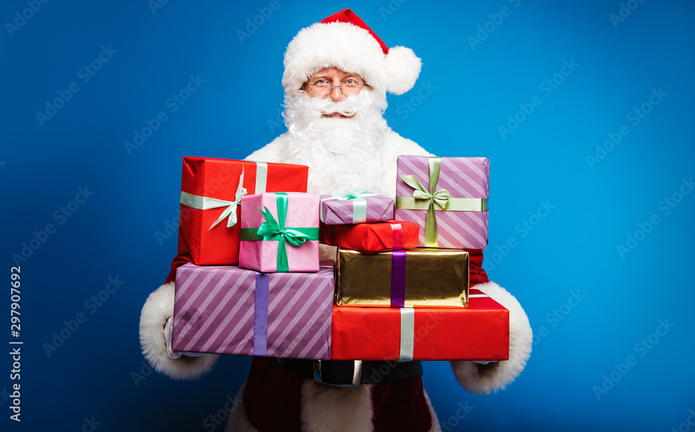 Everyone will be pleased. Authentic Santa Claus is holding a pile of multicolored presents in his hands, looking at the camera with a kind smile. Everyone will be pleased. Authentic Santa Claus is holding a pile of multicolored presents in his hands, looking at the camera with a kind smile.