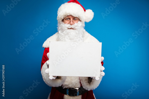 Read this. Close up photo of authentic Santa Claus in round-shaped glasses holding a blank postcard with a copyspace and showing it at the camera. © My Ocean studio