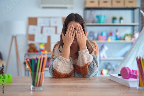 Young beautiful teacher woman wearing sweater and glasses sitting on desk at kindergarten rubbing eyes for fatigue and headache, sleepy and tired expression. Vision problem