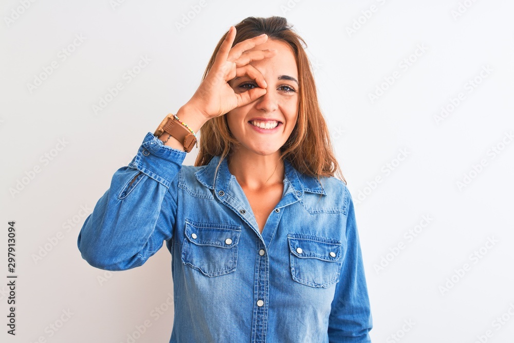 Young redhead woman wearing denim jacket standing over isolated background doing ok gesture with hand smiling, eye looking through fingers with happy face. Young redhead woman wearing denim jacket standing over isolated background doing ok gesture with hand smiling, eye looking through fingers with happy face.