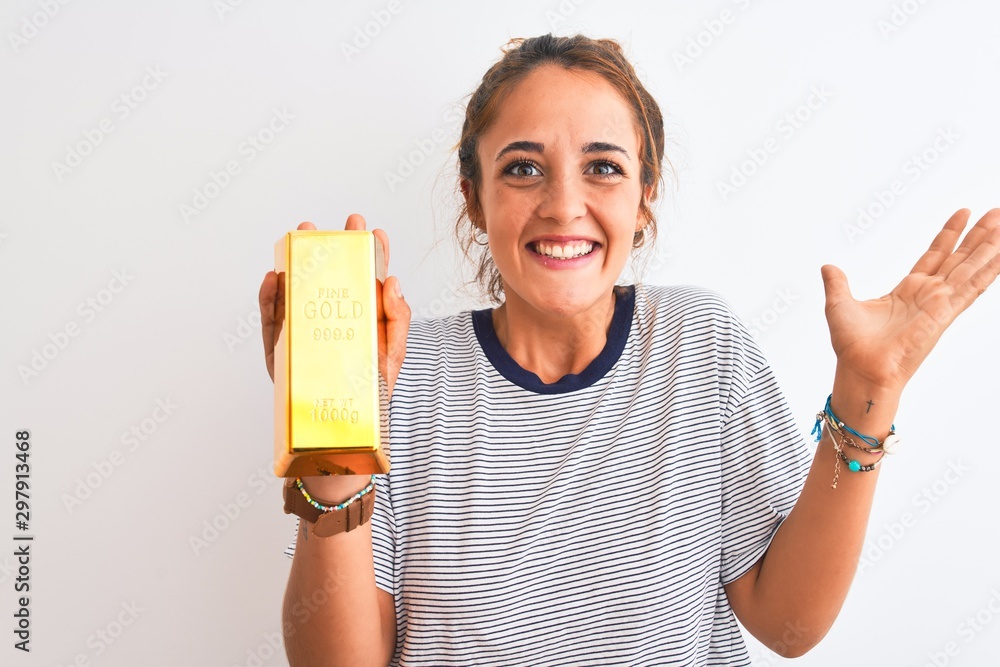 Young beautiful redhead woman holding gold ingot over isolated background very happy and excited, winner expression celebrating victory screaming with big smile and raised hands Young beautiful redhead woman holding gold ingot over isolated background very happy and excited, winner expression celebrating victory screaming with big smile and raised hands