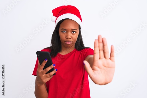 Young african american woman wearing christmas hat and using smartphone with open hand doing stop sign with serious and confident expression, defense gesture © Krakenimages.com