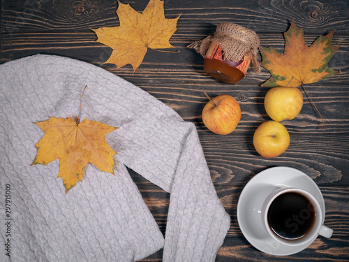 warm knitted sweater lies with autumn leaves, apples, honey and cup of coffee on wooden background 