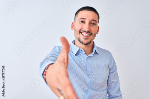 Young handsome business man standing over isolated background smiling friendly offering handshake as greeting and welcoming. Successful business.