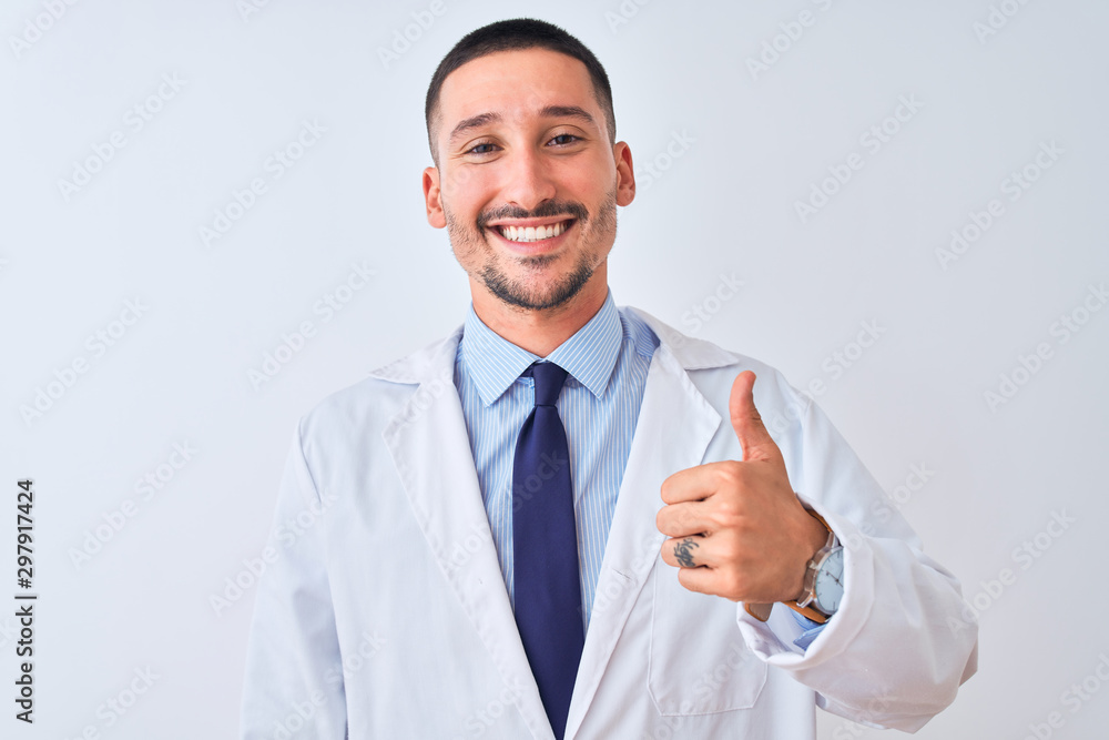 Young doctor man wearing white coat over isolated background doing happy thumbs up gesture with hand. Approving expression looking at the camera with showing success. Young doctor man wearing white coat over isolated background doing happy thumbs up gesture with hand. Approving expression looking at the camera with showing success.