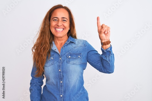 Middle age mature woman wearing denim jacket standing over white isolated background showing and pointing up with finger number one while smiling confident and happy.