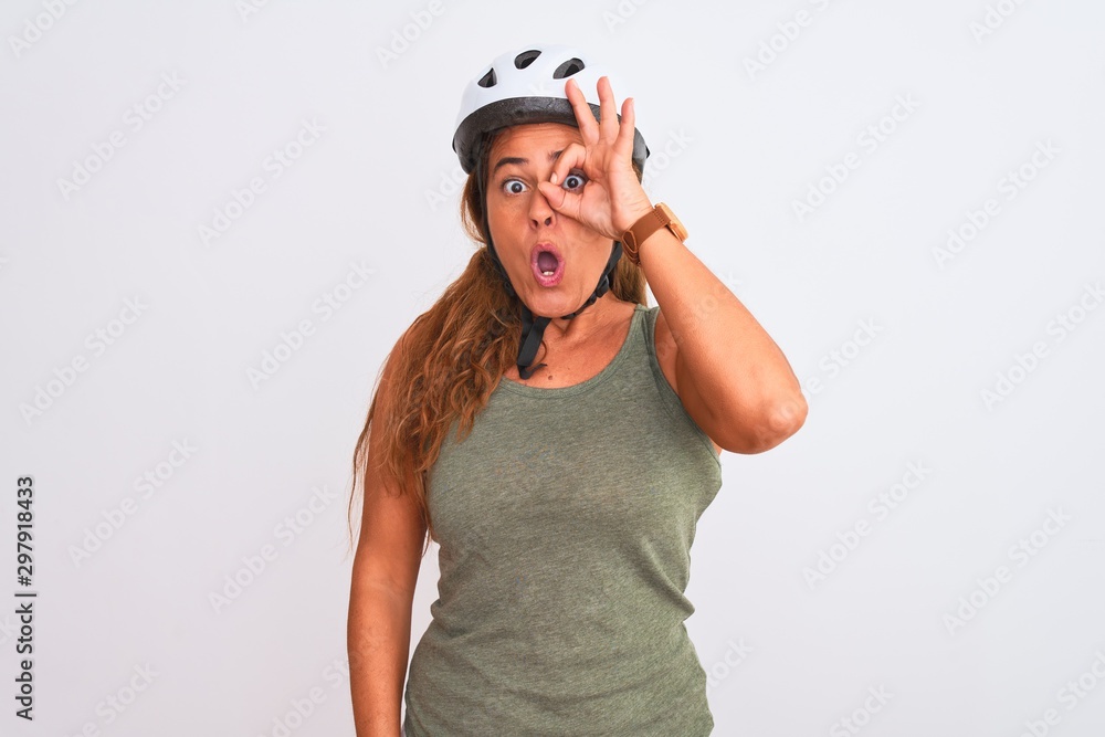 Fototapeta premium Middle age mature cyclist woman wearing safety helmet over isolated background doing ok gesture shocked with surprised face, eye looking through fingers. Unbelieving expression.