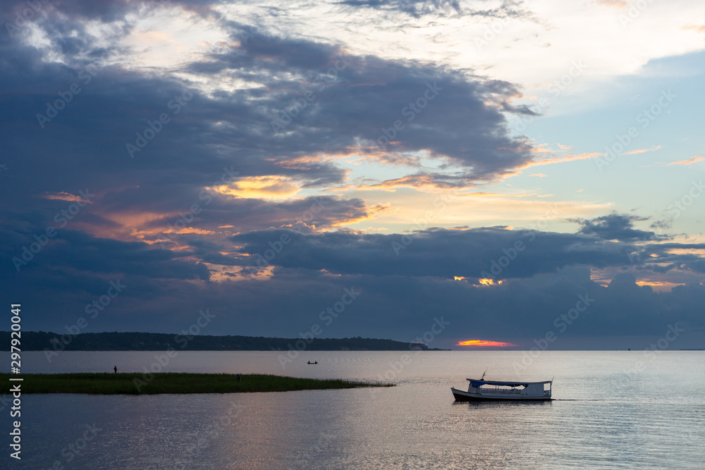 Wooden traditional boat sailing on Tapajos river at dusk with clouds and sunset in the background. Amazon, Brazil. Environment, climate change, travel, tourism and peace concept.