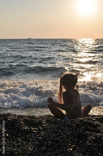 A little girl sits on the beach and looks at the waves in the rays of the sunset sea.