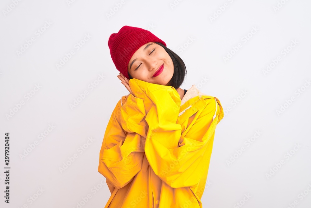 Young beautiful chinese woman wearing raincoat and wool cap over isolated white background sleeping tired dreaming and posing with hands together while smiling with closed eyes. Young beautiful chinese woman wearing raincoat and wool cap over isolated white background sleeping tired dreaming and posing with hands together while smiling with closed eyes.