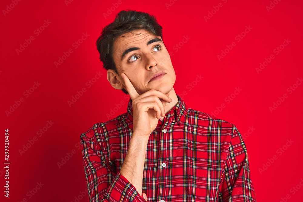 Teenager boy wearing red shirt standing over isolated background with hand on chin thinking about question, pensive expression. Smiling with thoughtful face. Doubt concept. Teenager boy wearing red shirt standing over isolated background with hand on chin thinking about question, pensive expression. Smiling with thoughtful face. Doubt concept.