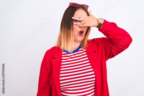 Young beautiful woman wearing striped t-shirt and jacket over isolated white background peeking in shock covering face and eyes with hand, looking through fingers with embarrassed expression.