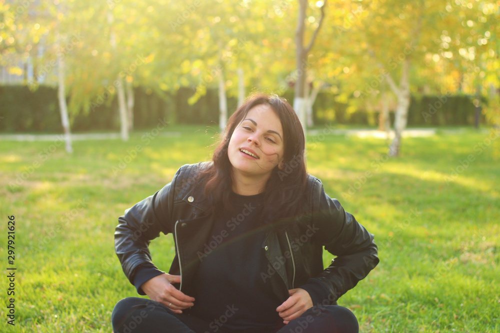 white girl with long dark hair is sitting in a leather jacket on a background of autumn yellow trees and grass with fallen leaves white girl with long dark hair is sitting in a leather jacket on a background of autumn yellow trees and grass with fallen leaves