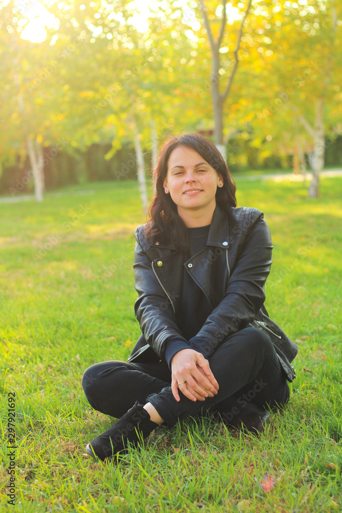 white girl with long dark hair is sitting in a leather jacket on a background of autumn yellow trees and grass with fallen leaves white girl with long dark hair is sitting in a leather jacket on a background of autumn yellow trees and grass with fallen leaves